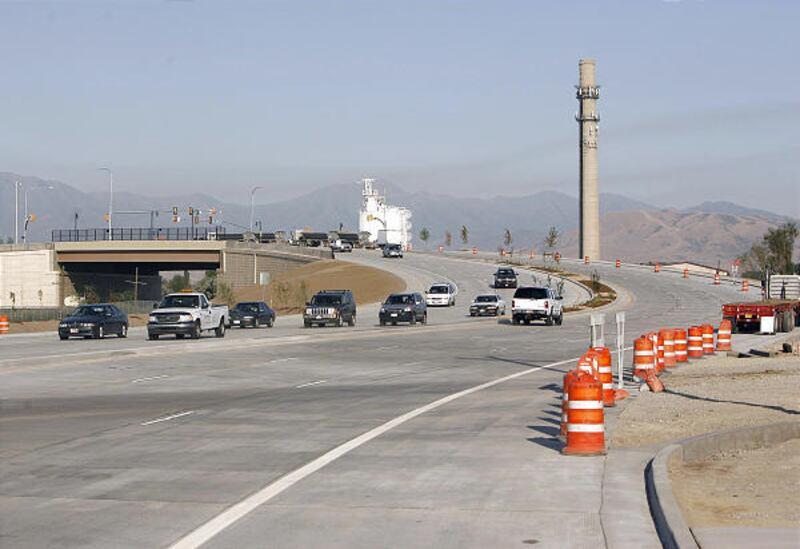 Wide view of Pioneer Crossing (SR-145) in Lehi, Utah with multiple traffic lanes, cars moving through a suburban area, nearby homes, and mountains in the background at sunset.