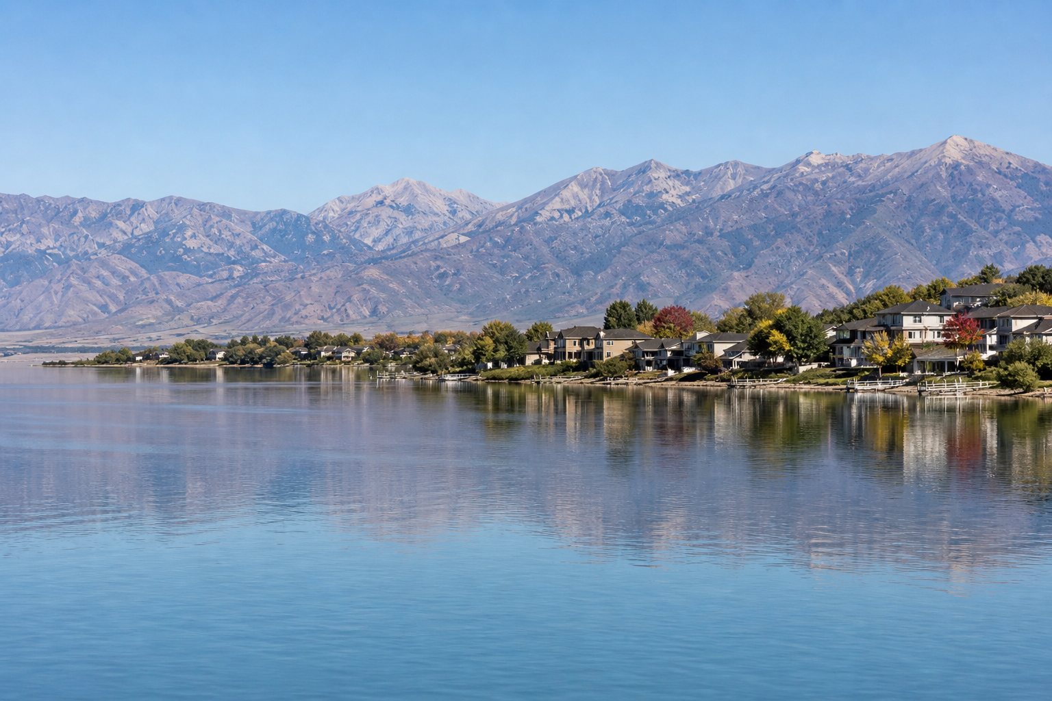 A wide, high-resolution landscape photo of Utah Lake with calm blue water in the foreground reflecting the shoreline. Along the right side of the lake, a row of suburban homes with docks and trees lin